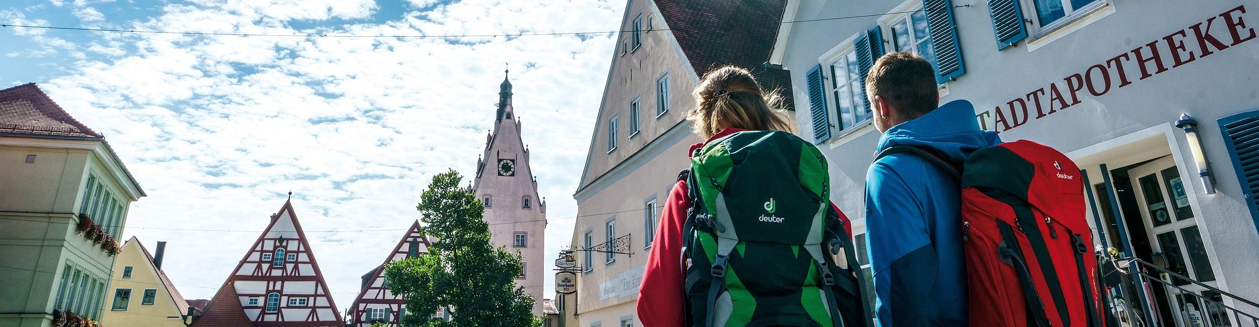 Wandern in der Drei-Stämme-Stadt -Monheim Zwei Personen mit Rucksäcken gehen an Gebäuden mit Giebeldächern und blauem Himmel mit Sonne vorbei.